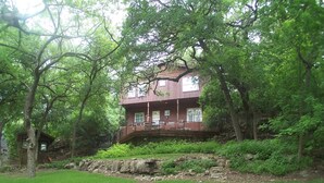 A two-story house with a balcony, surrounded by lush greenery and a stone wall.