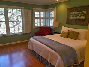 A bedroom with a large bed, two windows with shutters, a red chair, and a painting on the wall.