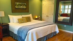 A neatly made bed with a white and gray bedspread, a nightstand with a lamp, a painting on the wall, a wardrobe, and a mirror reflecting the room.