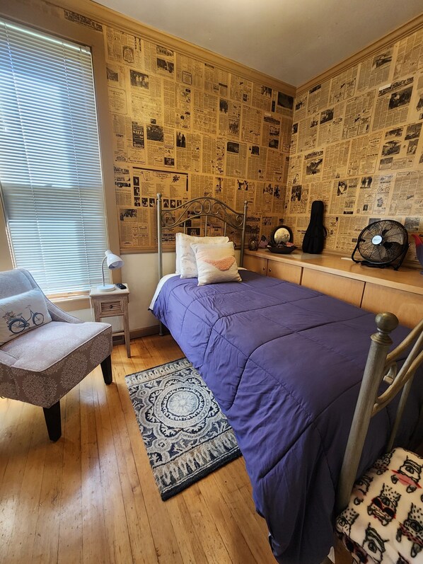 A bedroom with a bed, a chair, a nightstand, a fan, and a wall covered with newspaper clippings.