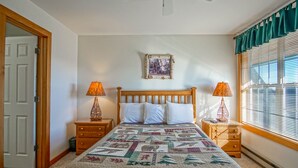 Un dormitorio con una cama de madera, dos mesitas de noche con lámparas, una colcha, un cuadro en la pared y una ventana con persianas.