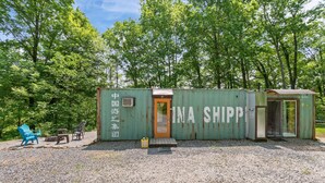 A rustic, green shipping container structure with a small porch and a door labeled "INA SHIPP."