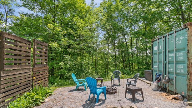 An outdoor seating area with wooden chairs and a fire pit, surrounded by a wooden fence and greenery. - A Shipping Container Experience (Dover)