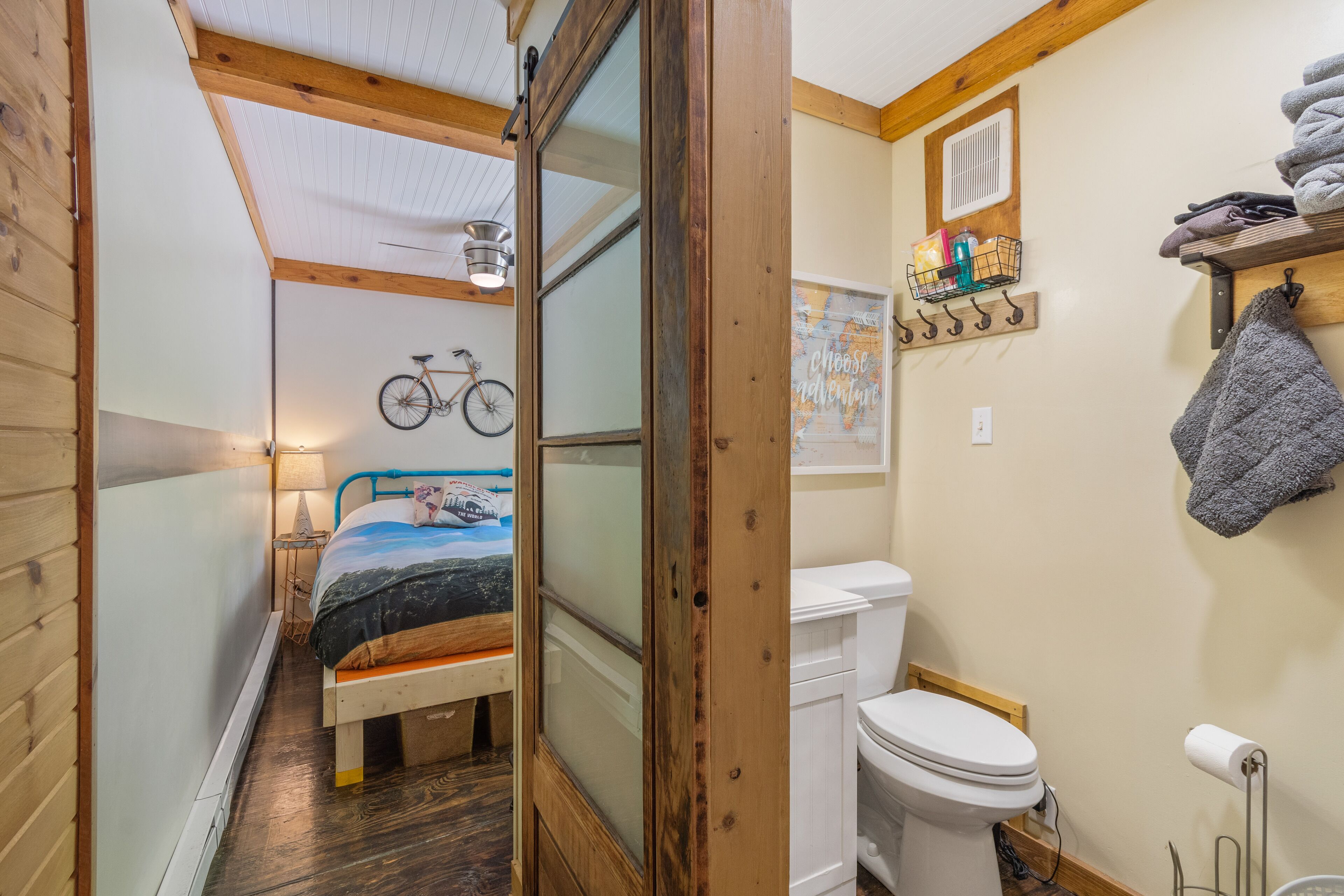 A narrow room with a bed, a bicycle, a toilet, and a shelf with towels.