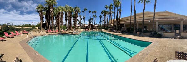 A swimming pool with clear blue water, surrounded by palm trees and red lounge chairs.