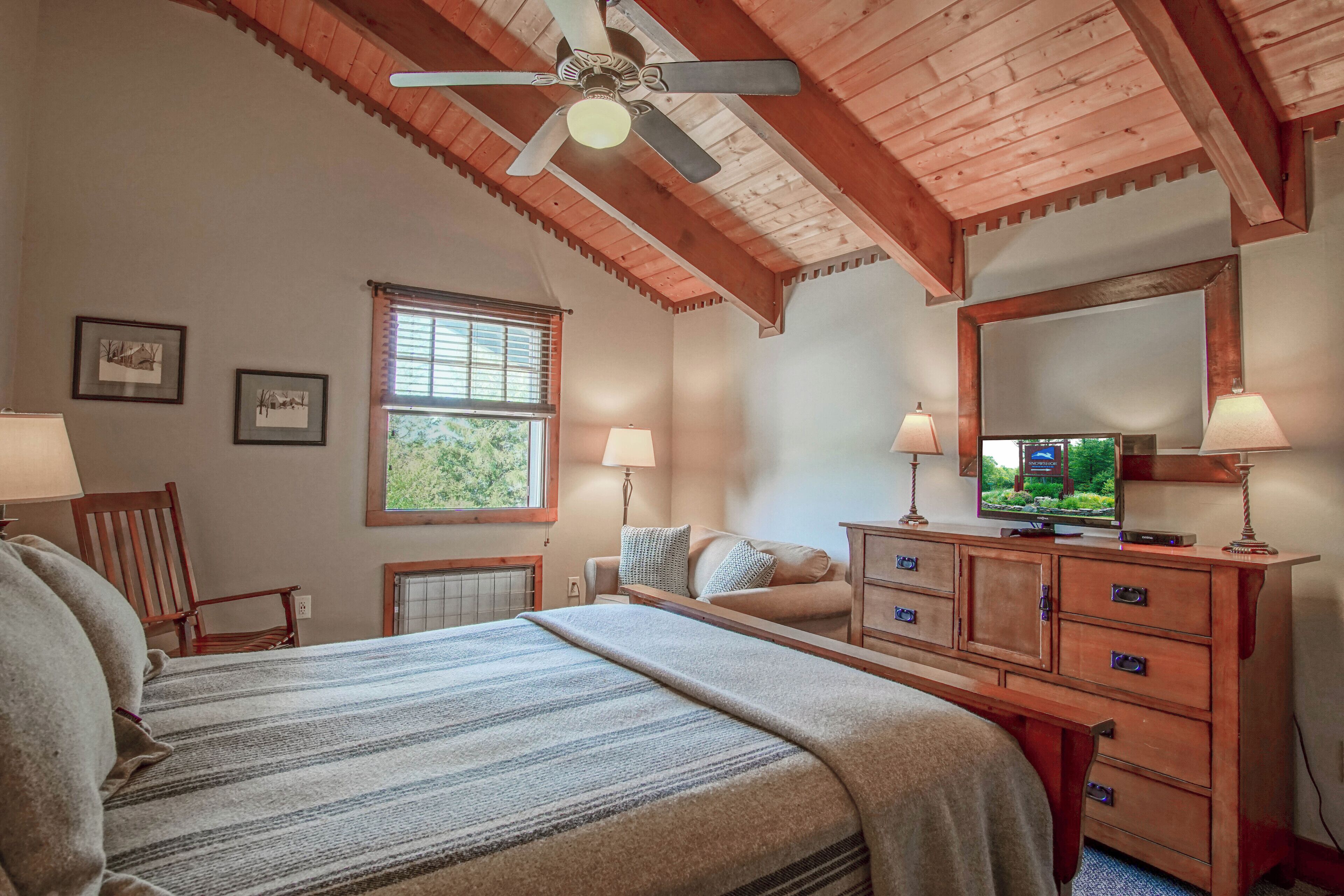 A bedroom with a bed, wooden dresser, ceiling fan, window, and wall-mounted TV.