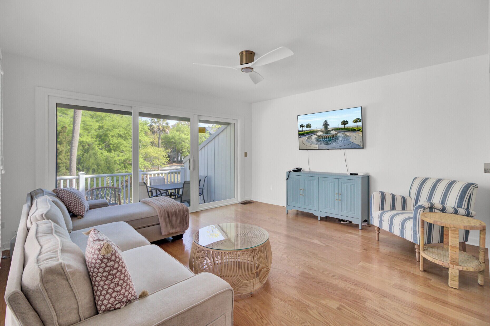 A spacious living room with a large sliding glass door leading to a deck, a ceiling fan, a flat-screen TV mounted on the wall, and a blue cabinet.