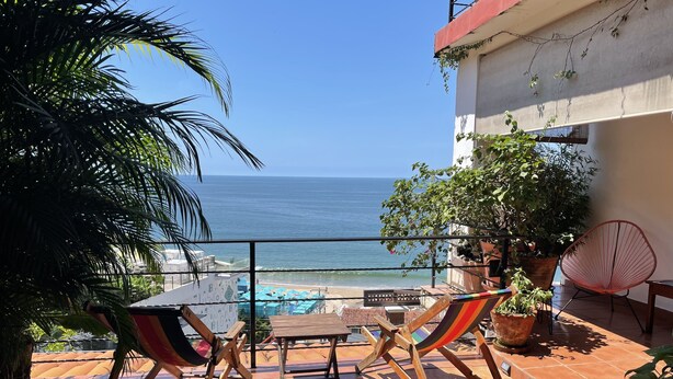 A terrace with a view of the sea, featuring a wooden table and chairs, potted plants, and a red chair. - Zona Romantica Area with Beautiful Bay View and Large Terrace (Puerto Vallarta)