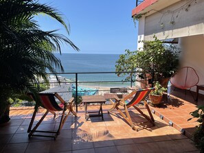 A terrace with a view of the sea, featuring a wooden table and chairs, potted plants, and a red chair.