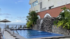 A swimming pool with a stone wall and a waterfall feature, surrounded by lounge chairs and a view of the ocean. - Zona Romantica Area with Beautiful Bay View and Large Terrace (Puerto Vallarta)