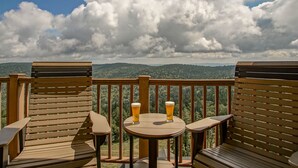 Una terraza de madera con dos sillas y una mesita pequeña, cada una con una cerveza. La terraza ofrece vistas a una cadena montañosa boscosa bajo un cielo parcialmente nublado.