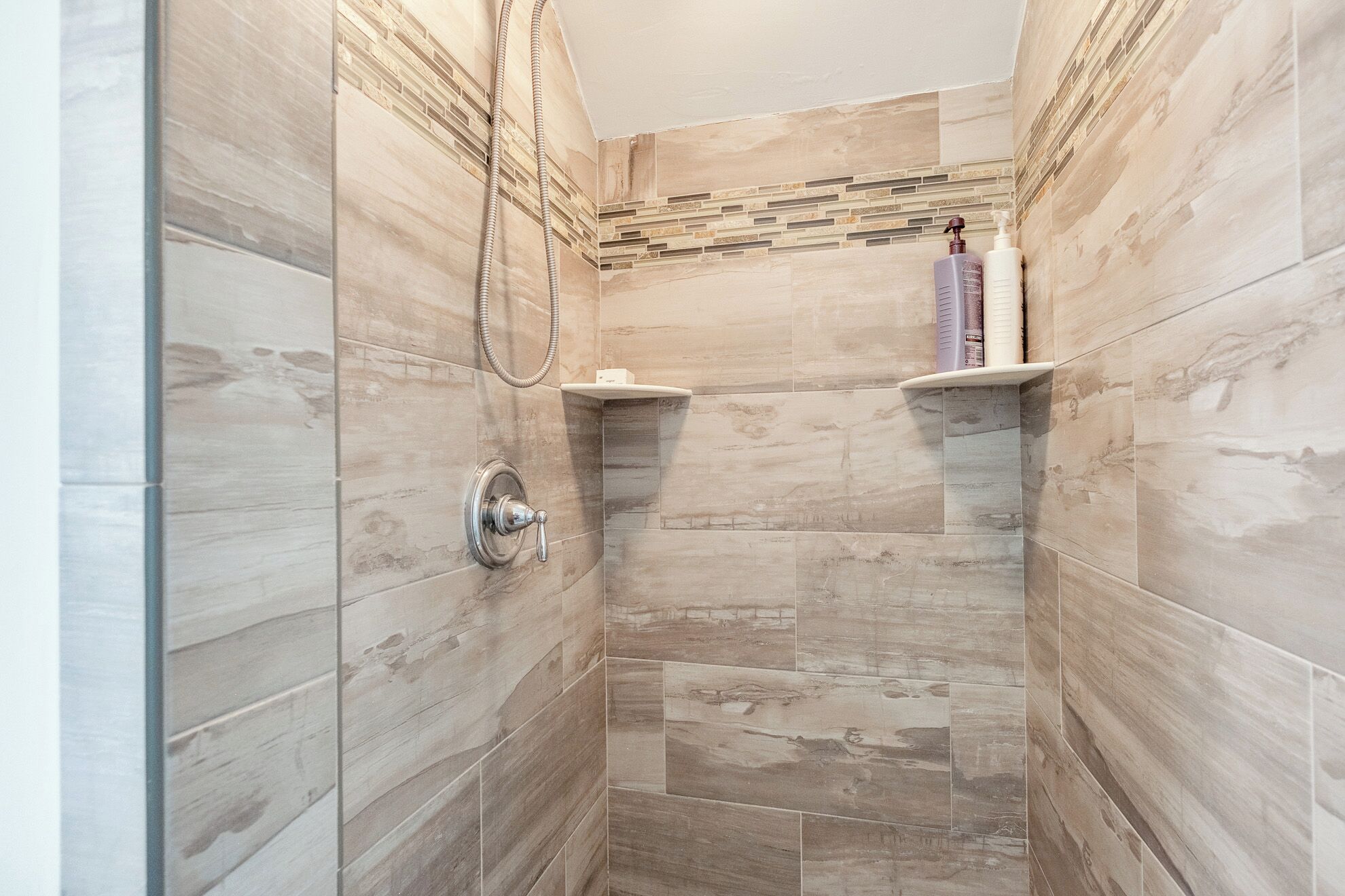 A shower area with a glass door, a handheld showerhead, and a shelf with toiletries.