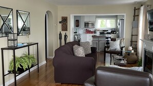 A spacious living room with a dark brown sofa, a leather armchair, and a small side table with plants. A kitchen with white cabinets and stainless steel appliances is visible in the background. - All Sports Lake / Golf, minutes from many players (Lapeer)