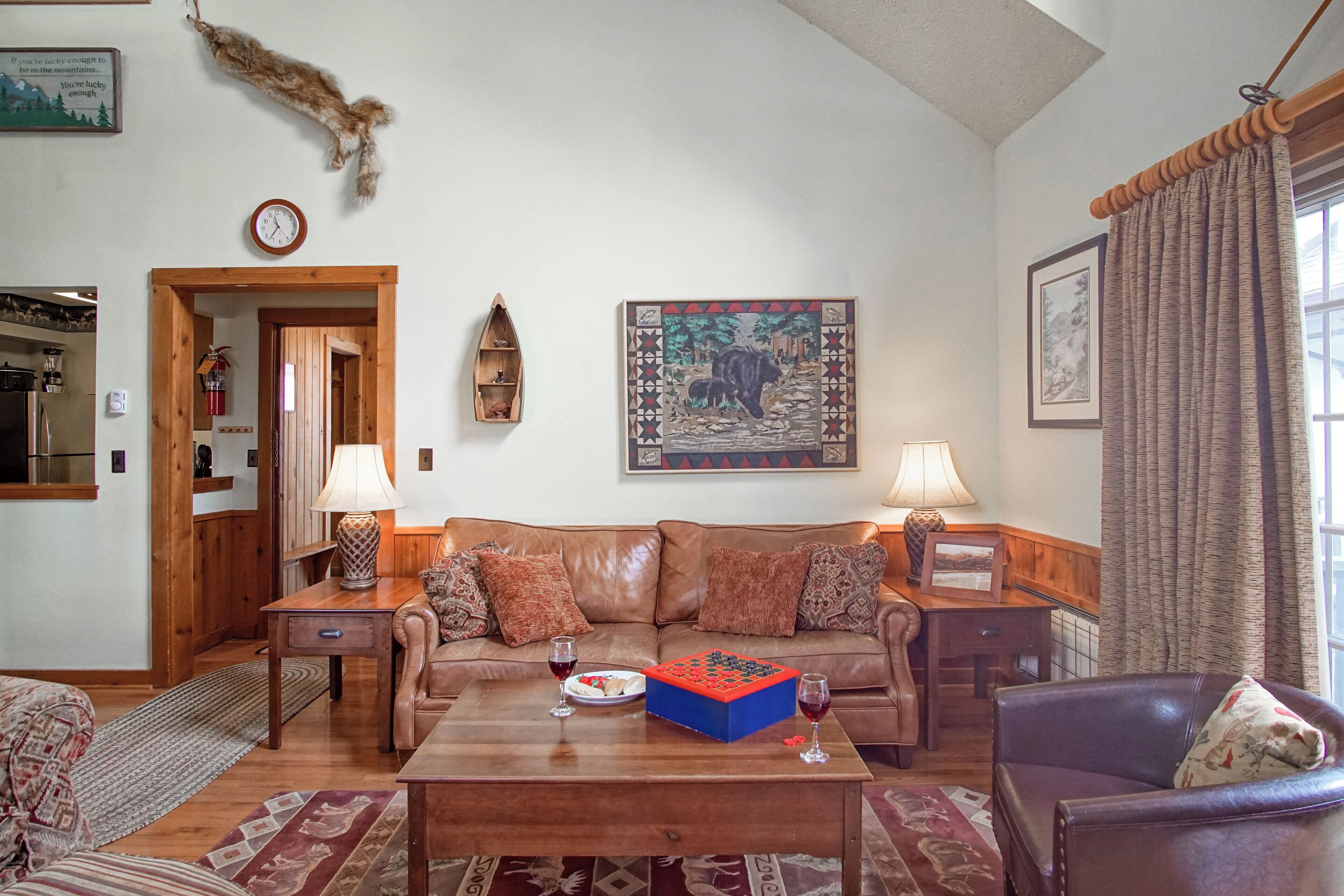A cozy living room with a brown leather sofa, a wooden coffee table, and a dining table with a plate of food and drinks. There are two lamps on side tables, a clock on the wall, and a painting above the sofa.