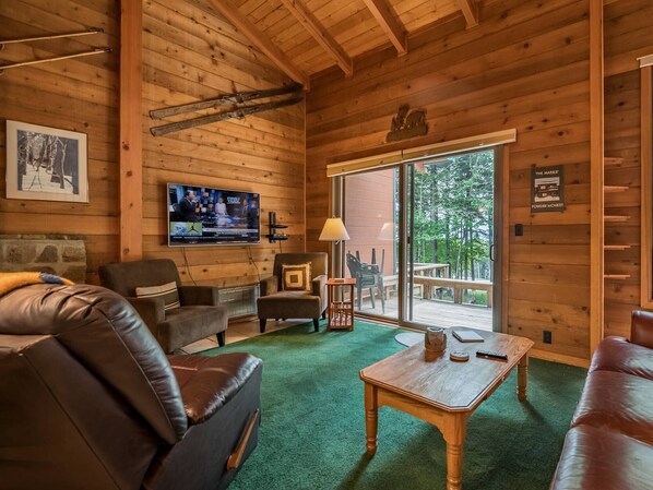 A cozy cabin interior with a wooden ceiling, a television mounted on the wall, a fireplace, and a large window offering a view of the outdoors.