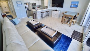 Una sala de estar moderna con un sofá, otomanas y una mesa de centro. Hay una zona de cocina con refrigerador, microondas y taburetes de bar. Un televisor está montado en la pared, y hay un cuadro de veleros.