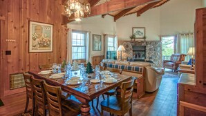 A wooden dining room with a large table set for six, surrounded by wooden chairs. There is a stone fireplace with a mantel, a sofa, and a chair in the background. - Whistlepunk #24: 4 Bedroom, 3 Bath:  Slope Side, Wood Fireplace. (Snowshoe)