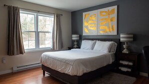 A bedroom with a bed, a window with curtains, a nightstand with a lamp, and a wall with abstract art. - Urban Edition (Long Pond)
