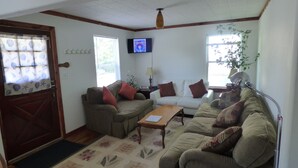 A living room with a wooden door, a television mounted on the wall, and a large window with a plant. - Cedar Haven Rolla - Relax in comfort close to downtown! (Rolla)