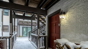 Un escalier en bois recouvert de neige mène à une porte rouge. À droite, on aperçoit une pile de bois de chauffage et, sur le mur, une lanterne.