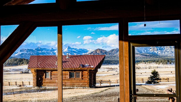 Une cabane en rondins surmontée d’un toit en métal, entourée d’un paysage enneigé et avec des montagnes en arrière-plan.
