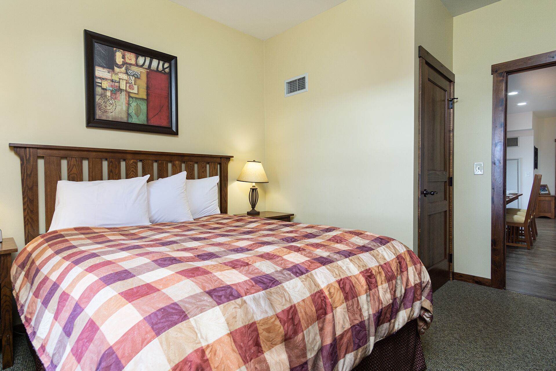 A hotel room with a large bed, a nightstand, a lamp, a painting on the wall, and a door leading to another room.