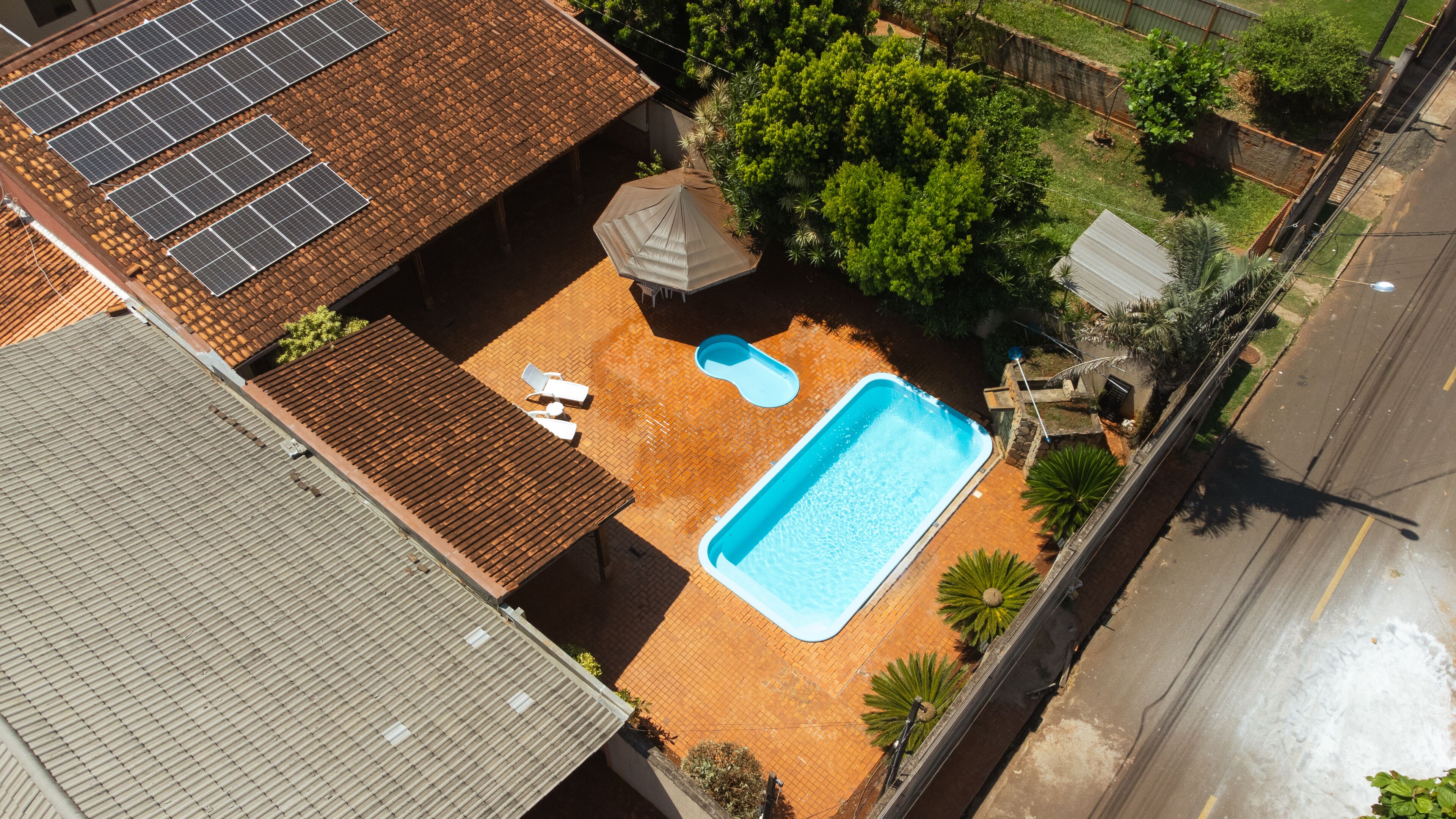 An aerial view of a residential area with a swimming pool, solar panels, and a fenced garden.