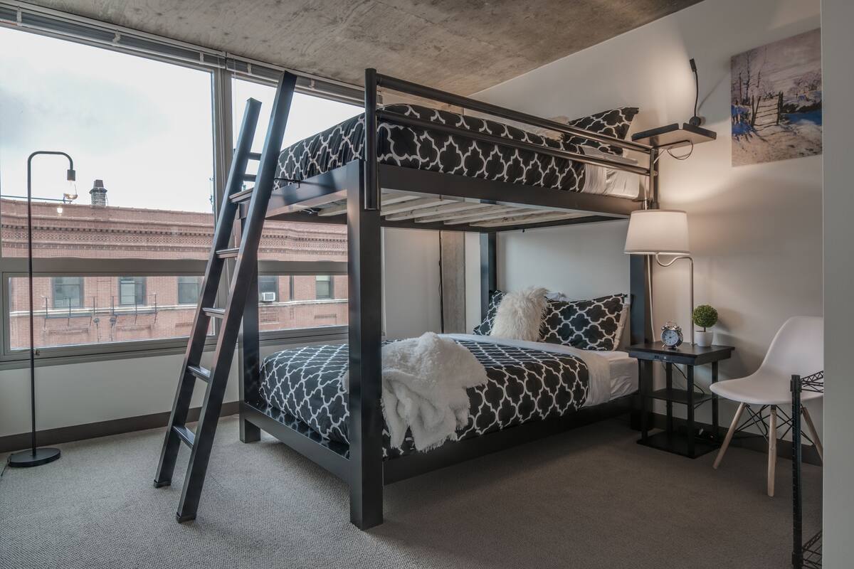 A bunk bed with patterned bedding, a desk with a lamp, a chair, and a view of buildings through a large window.