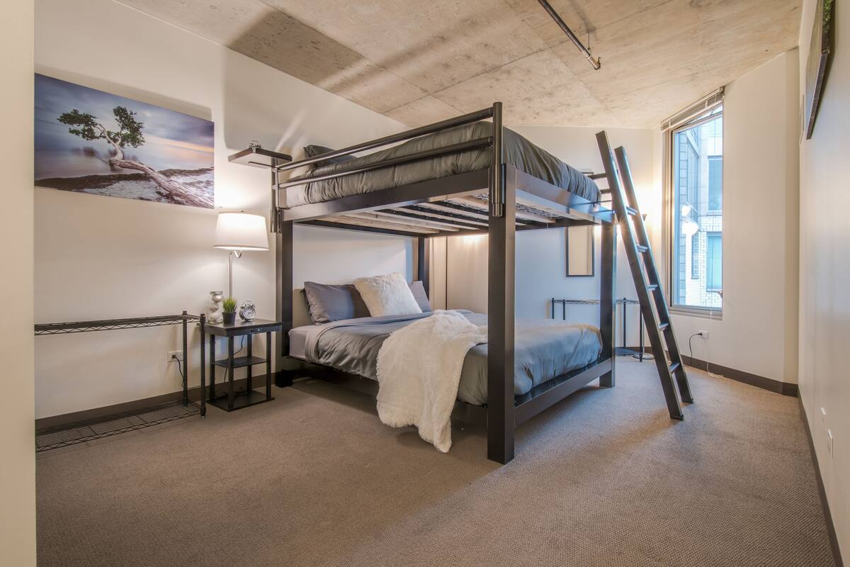 A bunk bed room with a ladder, a bedside table, a lamp, and a painting on the wall.