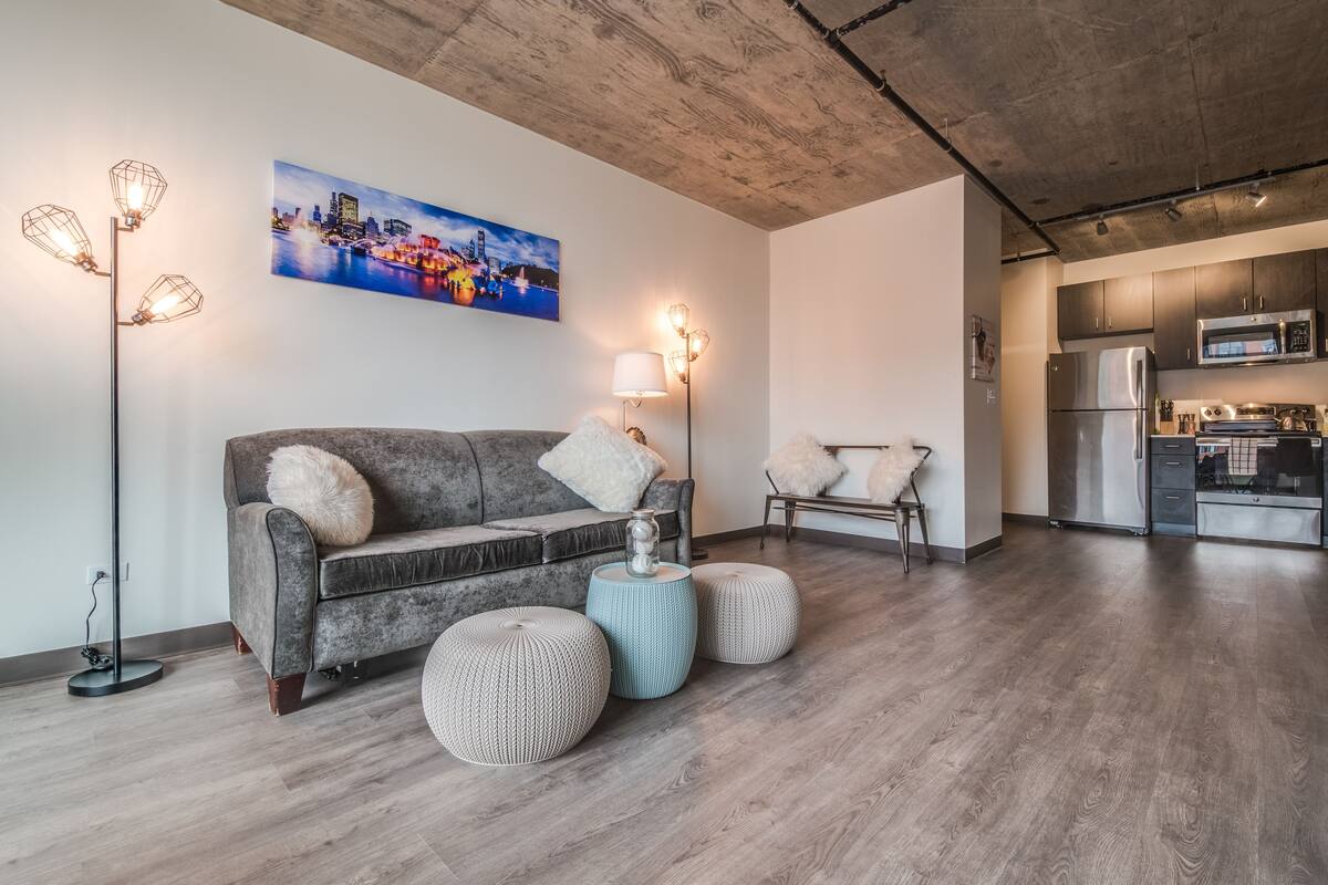 A modern living room with a grey sofa, round ottomans, a floor lamp, and a cityscape painting.