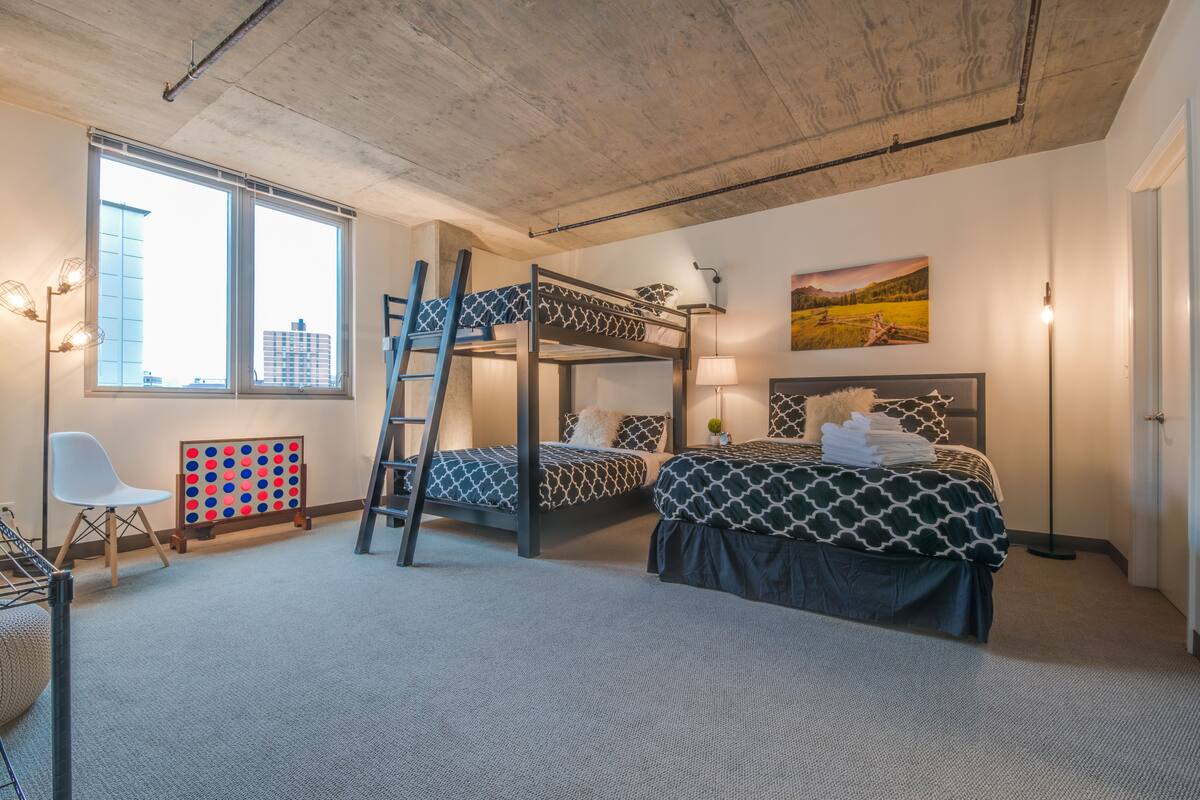 A dormitory room with bunk beds, a desk, a chair, a lamp, and a view of the city through large windows.