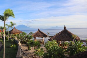 A beachside resort with thatched-roof structures, palm trees, and a view of the ocean and mountains. - Boga Segara Villa and Resto (Penida Island)