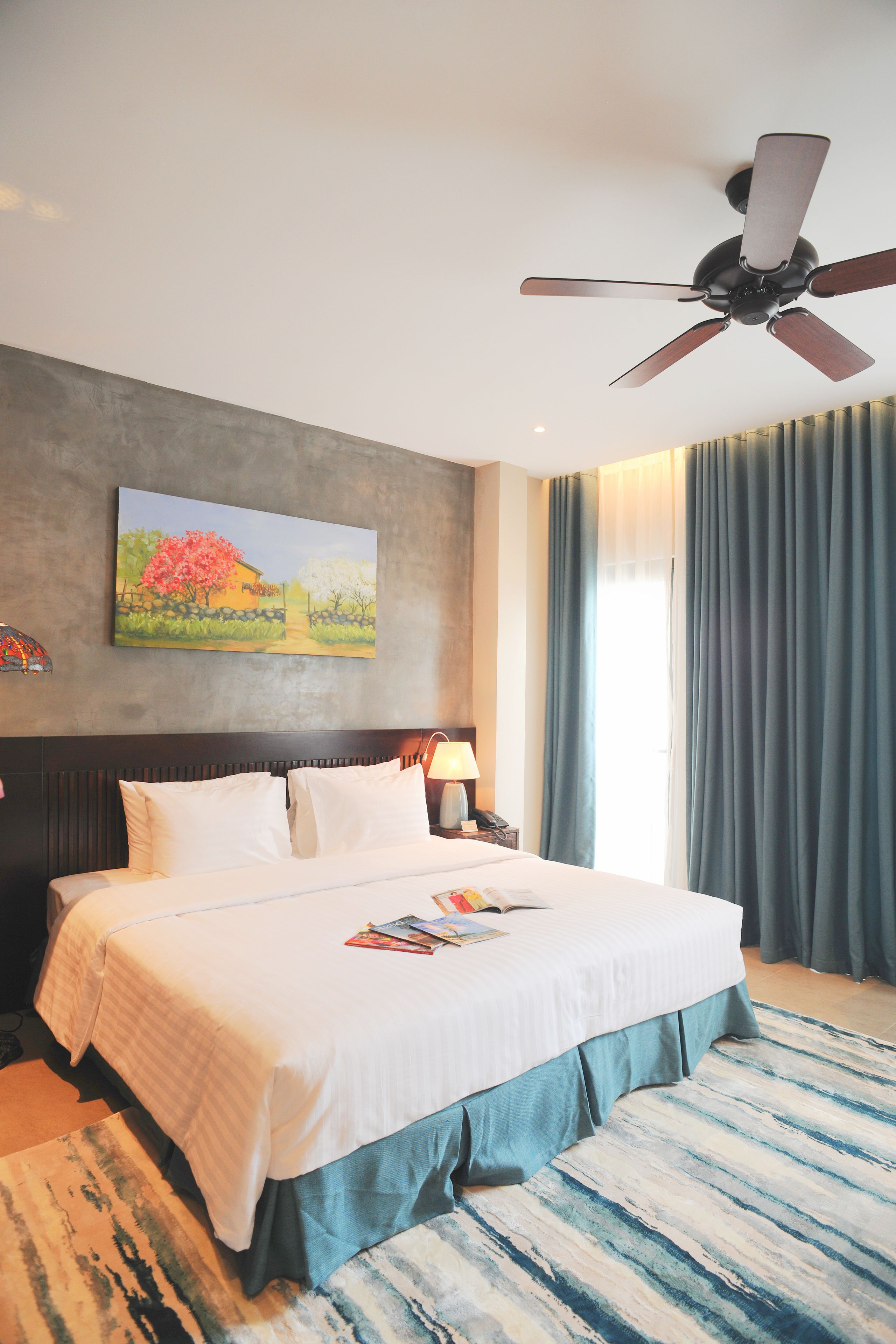 Habitación de hotel con una cama grande, ventilador de techo, un cuadro en la pared y una ventana con cortinas.