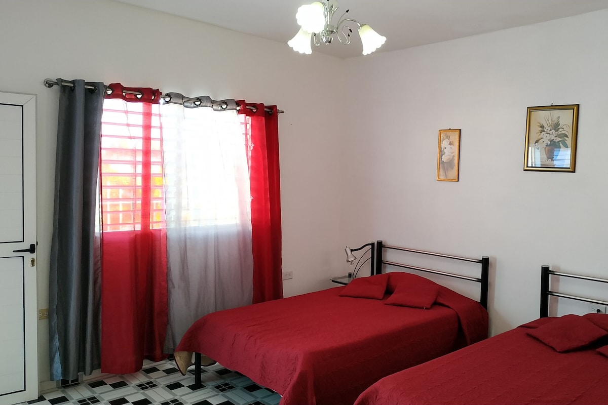 A bedroom with two single beds, a window with red and grey curtains, a ceiling fan, and three framed pictures on the wall.