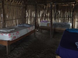 A rustic room with two beds, one with a floral mattress and the other with a patterned one. The room has a straw-like wall and a wooden floor.