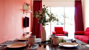 A dining area with a table set for two, a vase with green foliage, and a red sofa in the background. - PLAY Residence at Cameron Jaya (Tanah Rata)