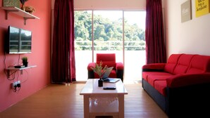 A living room with a red sofa, a red armchair, a coffee table with a laptop and a vase of flowers, a TV on a shelf, and a window with curtains. - PLAY Residence at Cameron Jaya (Tanah Rata)