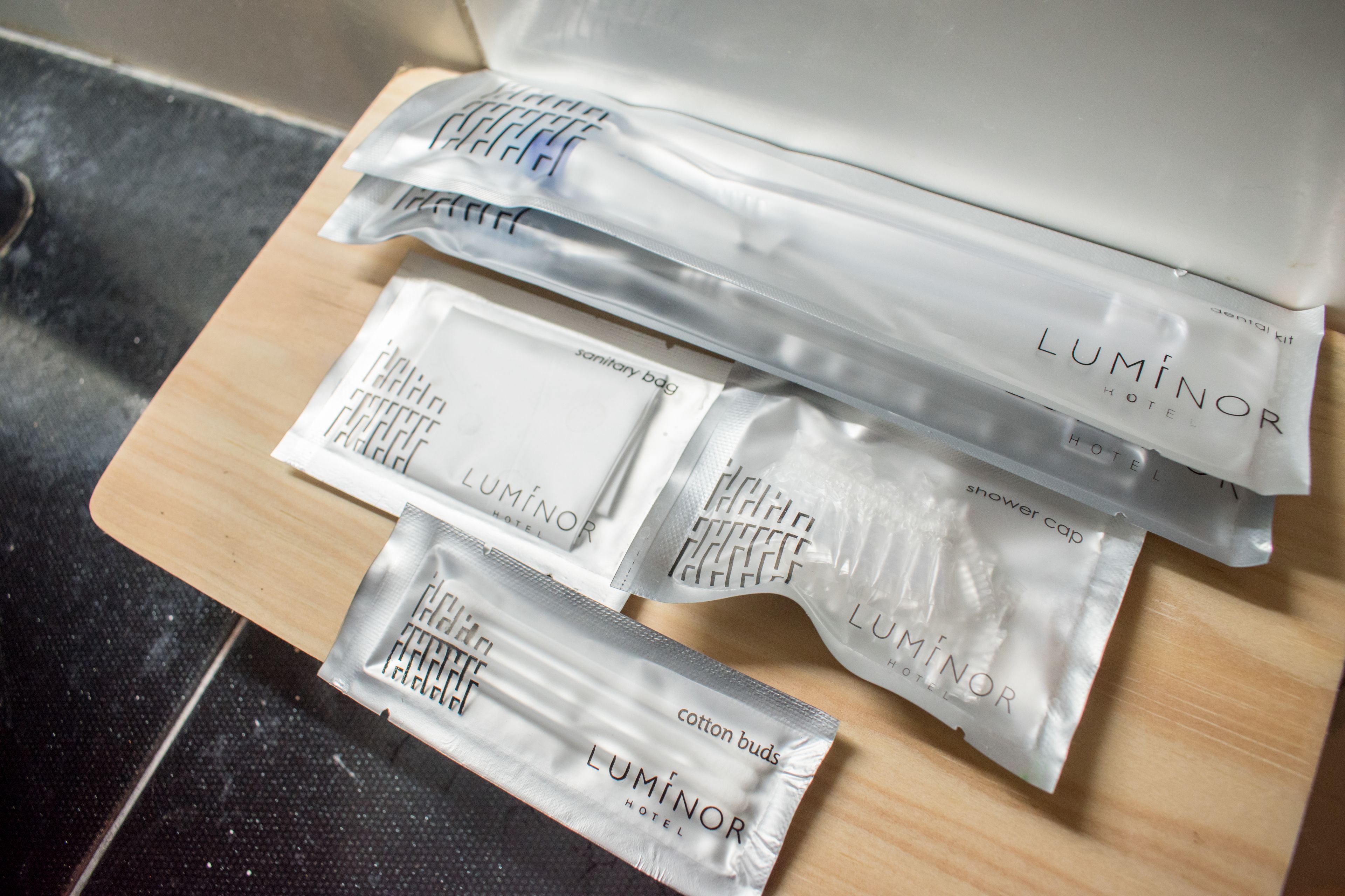 A wooden surface with several Luminor Hotel branded toiletry bags, including cotton buds and a shower cap.