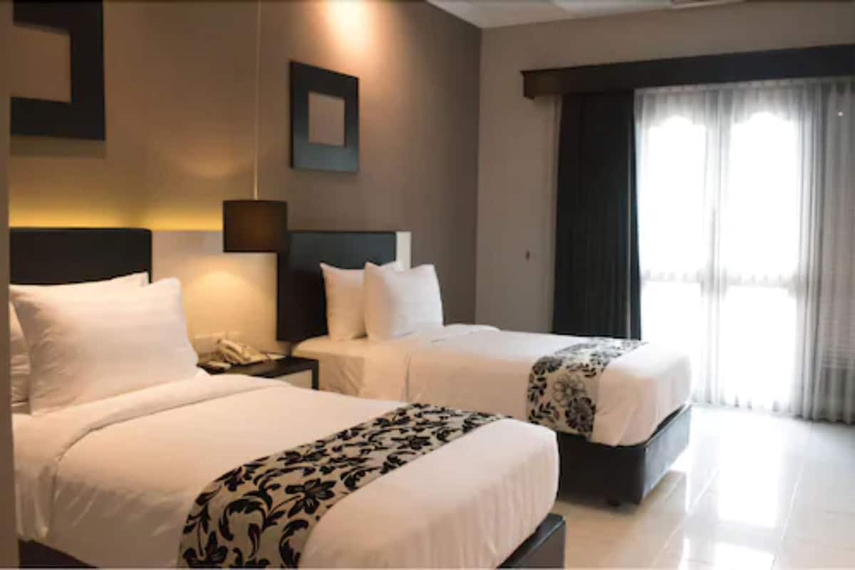 Habitación de hotel con dos camas, una mesita de noche con teléfono, una lámpara de pared y un ventanal con cortinas.