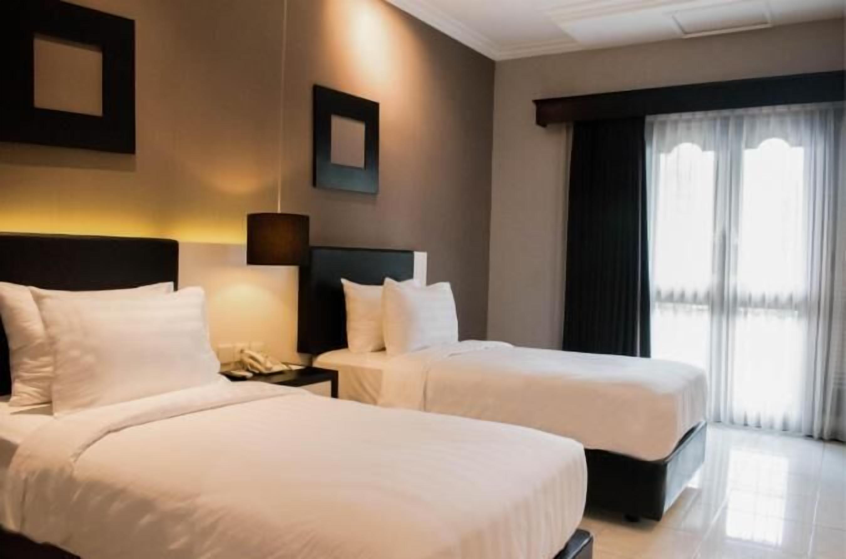 Habitación de hotel con dos camas, una mesita de noche, una lámpara y una ventana con cortinas.