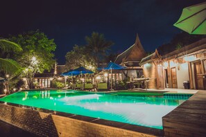 Un área de piscina nocturna con muelle de madera, sillones de descanso y sombrillas.