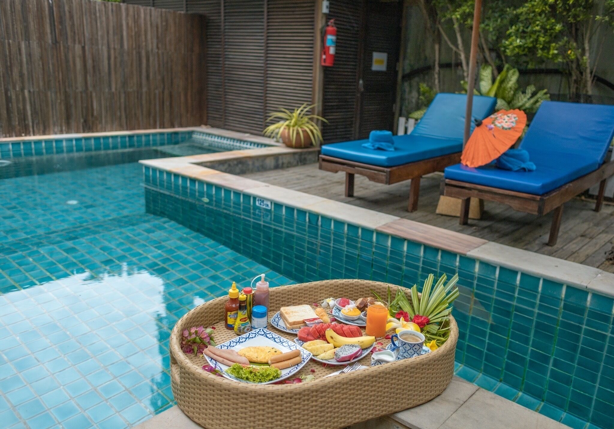 Une table de petit-déjeuner installée au bord de la piscine, avec un plateau en osier contenant de la nourriture et des boissons, deux chaises longues dotées de coussins bleus, et une clôture en bois en arrière-plan.