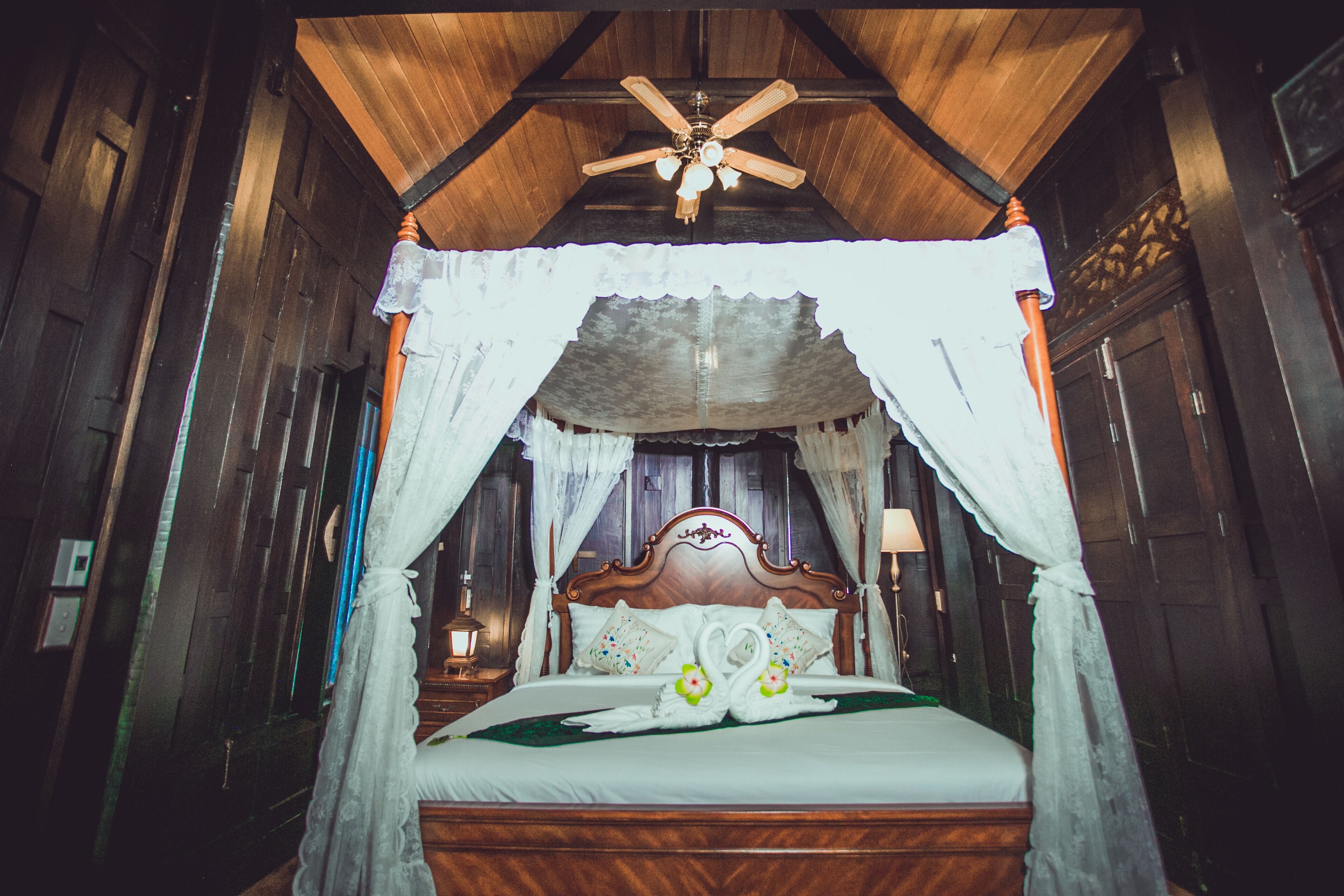 A wooden four-poster bed with a canopy, a ceiling fan, and a nightstand with a lamp.