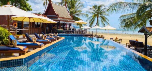 Una piscina con sillones y sombrillas, una playa al fondo y un edificio tradicional con techo inclinado.