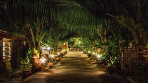 A pathway lined with potted plants and illuminated by soft lights, leading to a central area with a wooden bench and a hanging light fixture. - Sasitara   Thai  villas (Koh Samui)