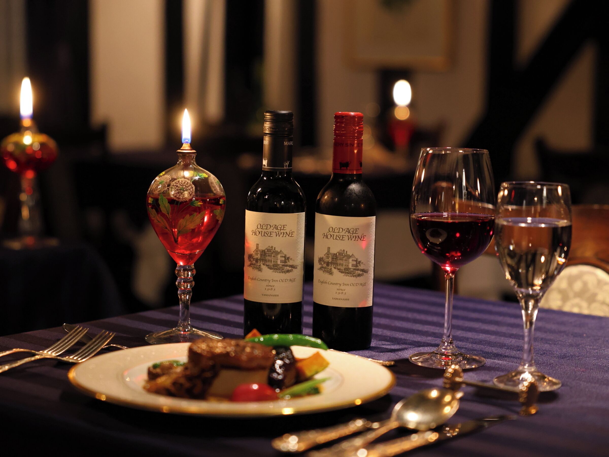 A dining table with a plate of food, two wine bottles, and three wine glasses.