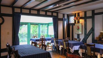 A traditional inn with exposed wooden beams, multiple dining tables with striped tablecloths, and a view of greenery outside.