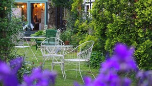 Un jardín con sillas y una mesa de metal blanco, rodeado de abundante follaje y flores moradas.