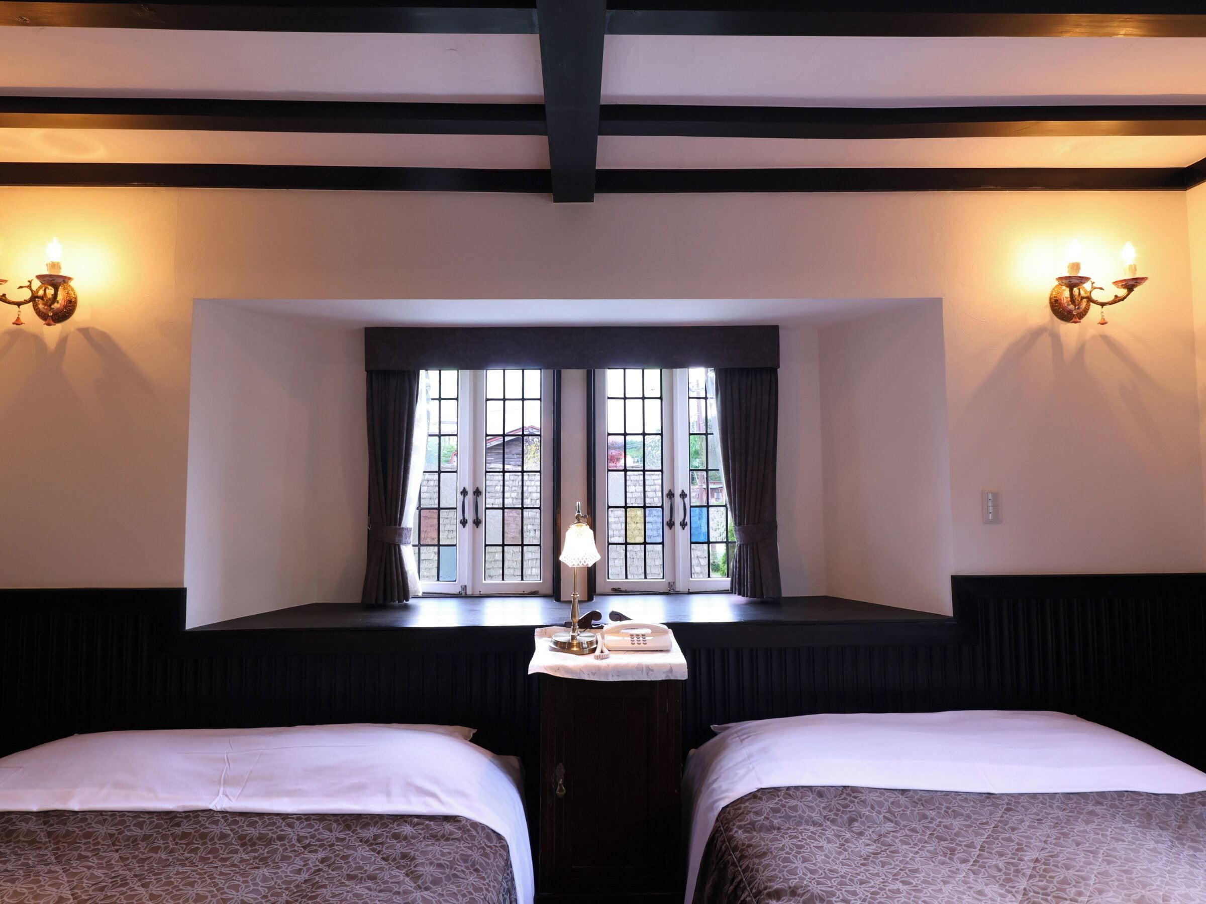 Habitación de hotel con dos camas, ventana con cortinas, mesita de noche con lámpara y apliques de pared.