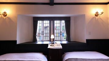 Habitación de hotel con dos camas, ventana con cortinas, mesita de noche con lámpara y apliques de pared.